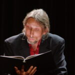 Rainer Rudloff im schwarzen Anzug vor schwarzem Hintergrund liest aus einem Text
