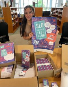 Eine Mitarbeiterin der Stadtbibliothek mit Plakat und anderen Materialien zum Sommerleseclub